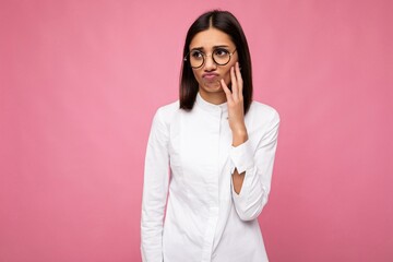 Photo shot of attractive upset sad dissatisfied young brunette female person wearing white shirt and stylish optical glasses isolated over pink background looking to the side