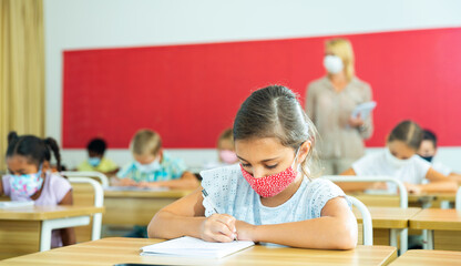 Young girl in protective mask is sitting at the desk in the classroom