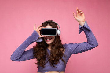 Photo of beautiful happy young brunette curly woman wearing purple crop top isolated over pink background wall holding and suggesting mobile phone with empty display and bluetooth earphones