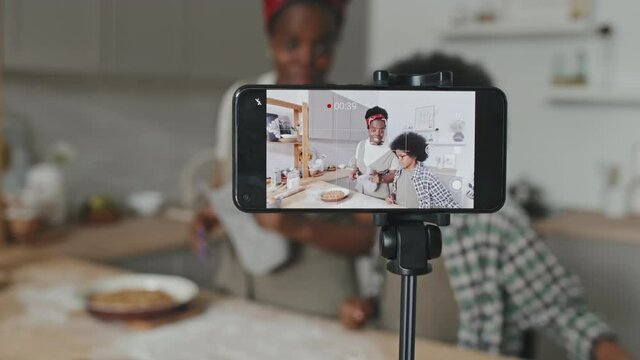 African American mother and son recording video of themselves on smartphone telling how to cook delicious homemade apple pie standing by table in cozy kitchen