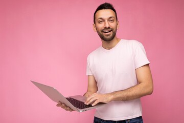 Handsome smiling brunet man holding laptop computer looking at camera in t-shirt on isolated pink background