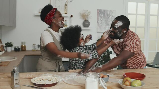 Medium slowmo shot of joyful African-American family of three having fun with flour while cooking together standing by kitchen table