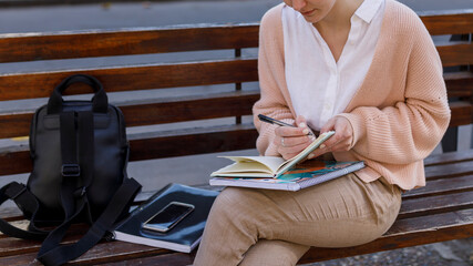 Young woman writing notes in a notebook, sitting on a city street bench. Studying or working outdoors.