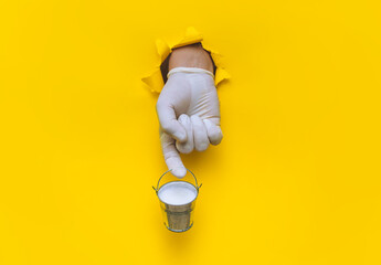 A right hand in a white medical glove holds a small iron bucket on the index finger with fresh milk. Farm hygiene and pasteurization concept. Yellow paper background with torn hole and copy space.