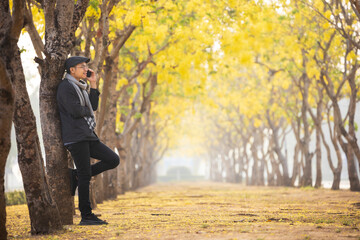 Asian man wearing sweater using mobile phone while leaning against yellow leaves ginkgo tree in the park in autumn season