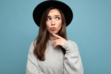 Photo of young thoughtful attractive pretty brunette woman with sincere emotions wearing casual grey sweater and black hat isolated over blue background with free space and thinking