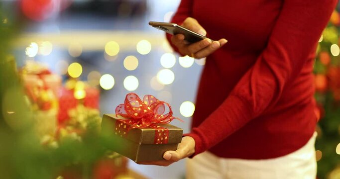 Woman in thick winter clothes using smartphone taking photos of red gift box in hand with bokeh lights in blur background. Concept of cheerful and alive of life in festival.