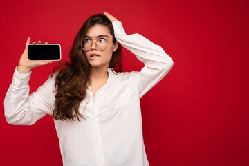 Naklejka premium photo of sexy beautiful young brunette woman wearing white shirt and optical glasses isolated over red background holding in hand and showing mobile phone with empty screen for cutout looking at