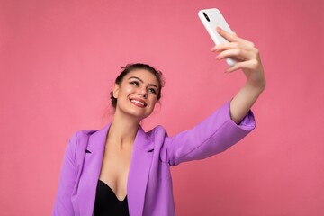 Attractive charming young happy woman holding and using mobile phone taking selfie wearing stylish clothes isolated over wall background