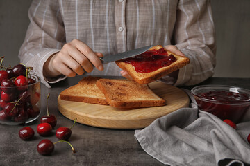 Woman spreading sweet cherry jam onto toast on dark background