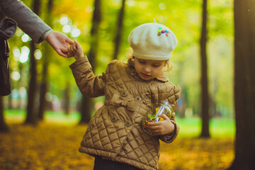 Little girl keeps glass jar filled with yellow and green leaves, flowers and seeds. Collected autumn treasures. Concept discovering and searching game for children. Daughter and mother hold hands