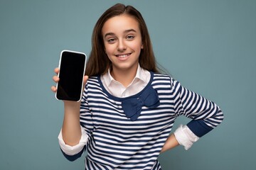 Pretty positive smiling girl wearing striped longsleeve standing isolated on blue background with copy space holding smartphone showing phone in hand with empty screen display for mockup pointing at