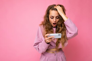 Photo of attractive crazy amazed surprised young woman wearing casual stylish clothes standing isolated over background with copy space holding and using mobile phone looking at camera