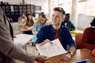 Happy college student takes exam paper from teacher in classroom.