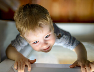 Close-up of happy baby boy standing at the window, reaching out with his hands to open the window pane. The child can not fly, close the windows. The concept of child safety and closed windows.