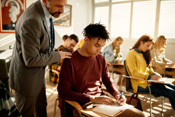 Black student writes an exam while professor is observing him in classroom.