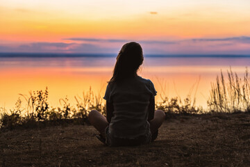 Fototapeta premium The silhouette of a little girl on the shore in front of a colorful orange sunset over the sea