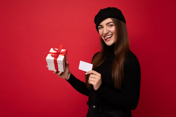 Fototapeta premium Beautiful happy young brunette woman wearing black sweater and hat isolated on red background holding credit card and gift box looking at camera