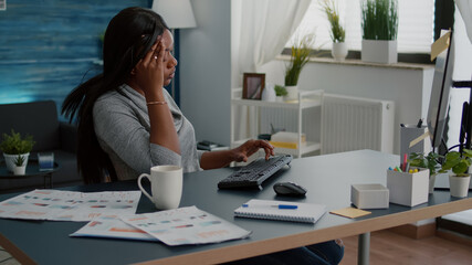 Tired distressed black student suffering headache sitting at desk table in living room searching medical treatment using computer. Worried sick upset young woman having migraine pain during lockdown