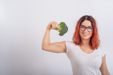 Caucasian woman holding broccoli and demonstrating biceps over white background. - obrazy, fototapety, plakaty