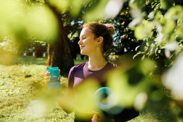 Portrait of smiling young sportive woman with closed eyes enjoying nature and sunny morning, sportswoman holding sports equipment for training, yoga mat and fitness water bottle. Yoga and Meditation