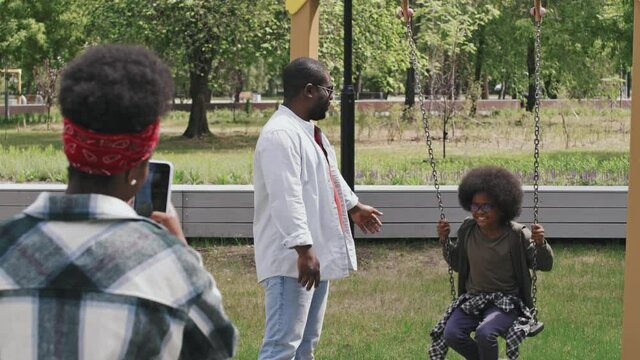 Slowmo shot of happy African American family sending summertime outdoors in park. Father swinging his son on chain swing while mother taking pictures of them on smartphone