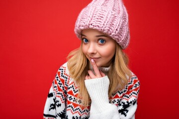 Photo of young beautiful blond lady with sincere emotions wearing pink knitted hat and winter pullover isolated over red background with empty space and showing shhh gesture with finger near lips. Don