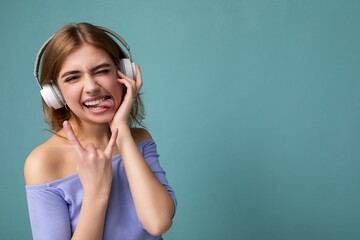 Photo of attractive emotional young blonde woman wearing blue crop top isolated on blue background wearing white wireless bluetooth headphones listening to cool music and having fun showing tongue and