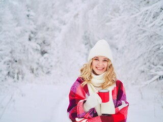 Obraz premium Beautiful happy smiling young woman in red plaid with cup of hot drink on cold snowy winter outdoors. Girl hands in gloves holding mug with hot cocoa, tea, coffee. Winter, Christmas time