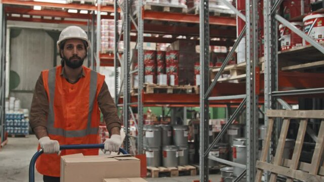 Slowmo tracking with tilt up of bearded male warehouse worker pushing cart and transporting items