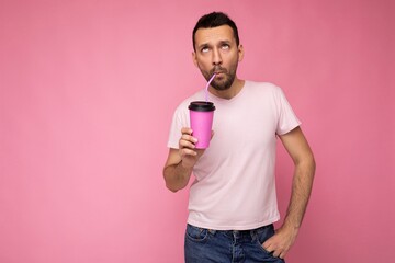 Handsome funny joyful young brunette unshaven male person with beard wearing white t-shirt isolated over pink backgroung wall holding paper cup drinking and enjoying