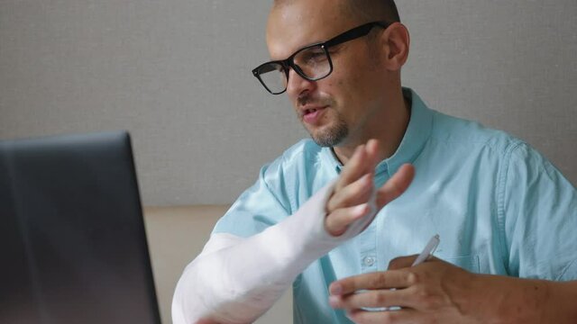Cheerful man in eyeglasses wearing blue shirt chatting online via laptop, showing his arm in plaster cast to webcam. Businessman working remotely from home after injury. Concept of communication