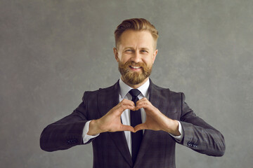 Portrait of handsome caucasian businessman in suit smiling showing heart shape as symbol of love. Cheerful man expresses his love standing on a gray background. Concept of romance and gratitude.