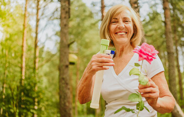 Fototapeta premium Elderly senior gardener farmer woman caring flowers in summer garden at countryside outdoors, sprays flowering plants using water pulverizer. Farming, gardening, agriculture, retired old age people
