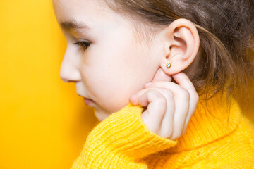 Ear piercing in a child - a girl shows an earring in her ear made of a medical alloy. Yellow...