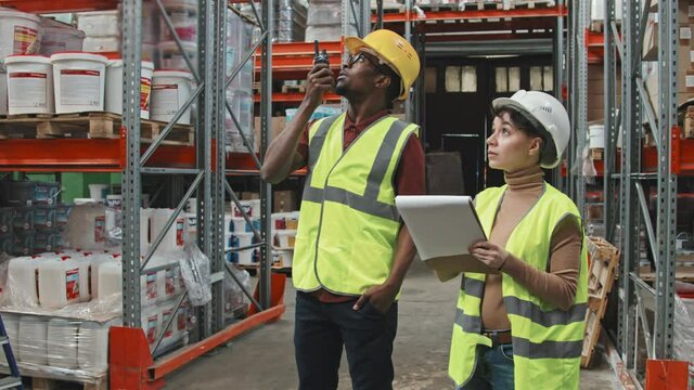 Handheld tracking with slowmo of Caucasian female warehouse supervisor in hard hat and safety vest writing on clipboard while her African-American male colleague talking into walkie-talkie