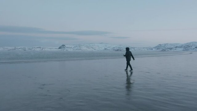A man goes along the wet sandy shore of the sea in a bay surrounded by snow cliffs. Slow motion