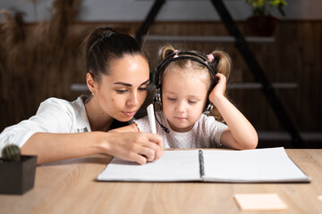 Happy young caucasian mom and handwriting small daughter in notebook prepare school home task together.