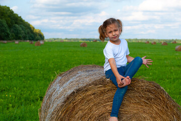 Naklejka na ściany i meble A little blonde girl in a white T-shirt sits on a haystack, winces and rubs the heel of her foot against the background of green grass and blue sky