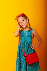 a fashionable blonde girl 8 years old European appearance with a phone in her hands and glasses on her head is talking on the phone. in a blue dress with a red bag over the shoulder. Place for text