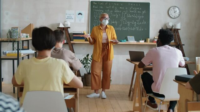 Tracking shot of female teacher in protective mask standing in front of multiethnic students in classroom and giving lesson while working during covid-19 pandemic