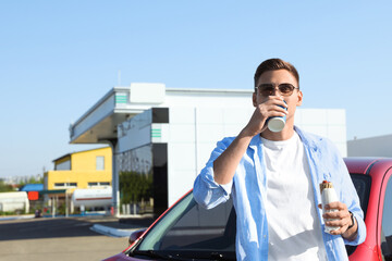 Young man with hot dog drinking coffee near car at gas station