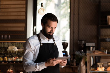 Attractive man in apron (barista or small business owner) stands in coffee house holding tablet in hands. Startup cafe owner concept	