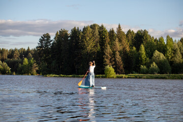 A young girl in a skirt rides a SUP board on a forest pond