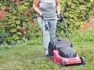 Senior woman mows the lawn using an electric pushing lawn mower.