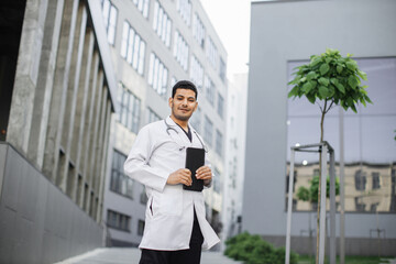 Male handsome Indian Asian doctor, wearing white coat, holding digital tablet in hands for working with patient's records, while standing outside modern clinic. People and technologies concept