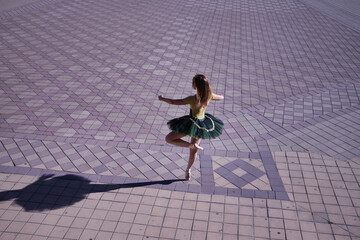 Adult female Hispanic classical ballet dancer in green and black tutu with coins, dancing in the middle of a plaza casting her shadow on the ground.