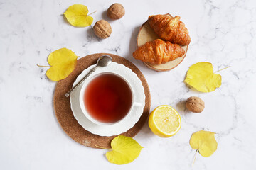 Cup of tea with falling yellow leaves, croissants, lemon and walnuts. Hello autumn. Tea break at home. Morning tea routine. English morning concept.  Thanksgiving holiday composition. Warm cozy home.