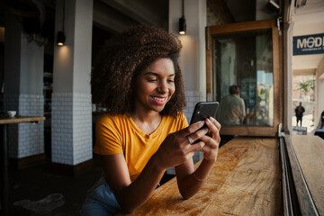 Smiling female customer texting off smartphone texting mates from smartphone sitting in trendy cafe 