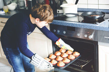 Happy blond school kid boy baking chocolate banana vanilla muffins in domestic kitchen, indoors....